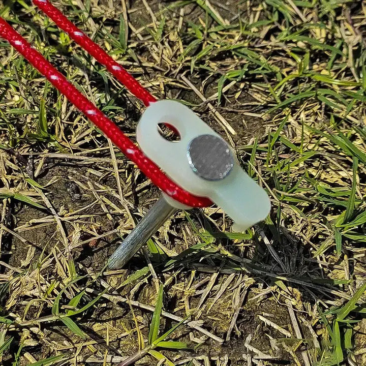 Luminous tent stake with red cord secured in grass, featuring high hardness steel and fluorescent plastic for outdoor camping.